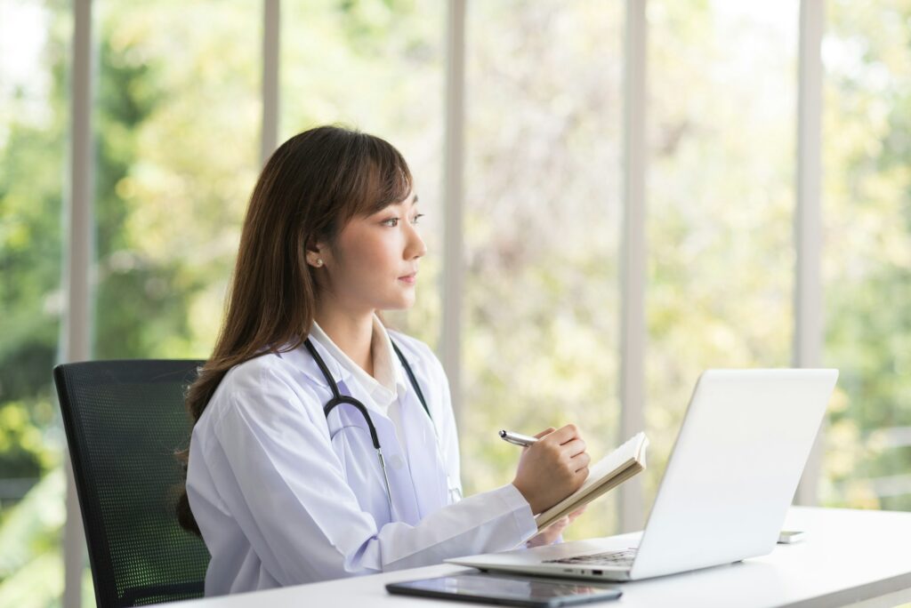 Asian doctor woman sitting and working with her laptop in modern office clinic room.